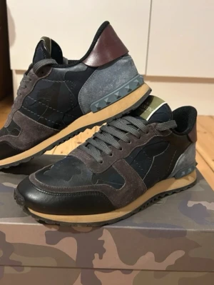 Valentino Garavni rock runner  - Valentino Garavani Rockrunner, strl 40  Äkta Valentino, köpta hos Kurt Geiger. Nypris £500. Läder och mocka med camomönster och nitar vid hälen.  Använda men i bra allmänt skick. Originalkartong och dustbags ingår.  Notera: • Liten reva i hälfodret på båda skorna • En liten reva på insidan Allt syns på bilderna och går att laga hos skomakare eller täckas med hälskydd. Jag kan fixa det om önsk  Fina, rena och redo att användas. Säljs på grund av garderobrensning. Skriv om du vill ha fler bilder. 