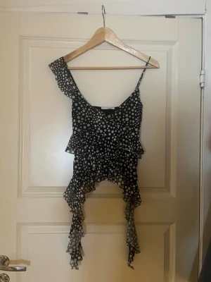 Prickig volangtop från Zara - Säljer en svart topp från Zara med vita prickar och volangdetaljer. Toppen har asymmetrisk design med en axelbandslös sida och en smal axelrem. Materialet är lätt och luftigt, perfekt för varma dagar. Volangerna ger toppen en cool och lekfull look. Oanvänd men prislappen är avklippt. 