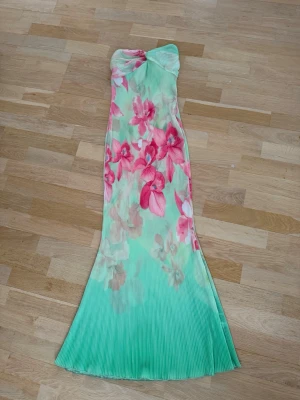 Mintgrön blommig långklänning - Superfin axelbandslös långklänning i mintgrönt med stora rosa och beige blommor. Klänningen har plisserad struktur och en snygg twistad detalj upptill. Perfekt för sommarens alla tillfällen! Genomskinlig