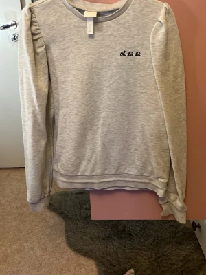 Ljusgrå sweatshirt från H&M med puffärm - Superskön ljusgrå sweatshirt från H&M i storlek S. Tröjan har puffärmar och ett litet broderat 'oh là là' på bröstet. Tillverkad i mjuk bomullsmix och har rund halsringning. Perfekt för en chill och trendig look.