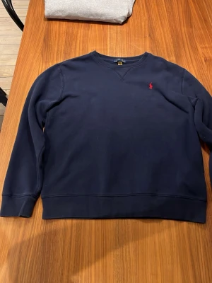 Mörkblå sweatshirt från Polo Ralph Lauren - Klassisk mörkblå sweatshirt från Polo Ralph Lauren i storlek M. Tröjan har rund halsringning, långa ärmar och den ikoniska röda broderade loggan på bröstet. Tillverkad i mjuk bomull för skön vardagsstil.