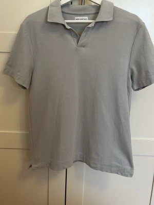 Ljusgrå piké från These Glory Days - Säljer en stilren ljusgrå pikétröja från These Glory Days i storlek medium. Klassisk krage, korta ärmar och knappar vid halsen. Tillverkad i mjuk bomull som känns skön mot huden. Perfekt för en clean och enkel look.