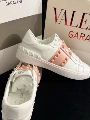 Valentino Rockstud sneakers vit/rosa - Säljer ett par vita Valentino Rockstud sneakers med rosa band och ikoniska nitar. Skorna har rund tå, platt sula och snörning. Tillverkade i skinn för en lyxig känsla och detaljerade med ljusrosa accenter och pyramidformade nitar runt hälen och ovandelen.