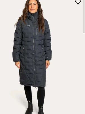 Svart lång dunjacka från uhip(ridjacka) - Svart dunjacka(snarare mörkgrå)  med huva och vita detaljer från uhip, nervikbara ärmar,två sidofickor med dragkedja och reflekterande ränder på ärmarna, skön luva och är även uppknäppbar där bak för att få en bra sits. Perfekt för kalla dagar.                                      Ny pris ca 3000kr