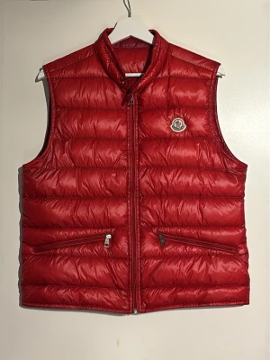 Röd dunväst från Moncler  - Säljer en röd dunväst från Moncler med klassisk quiltad design och hög krage. Västen har två dragkedjeförsedda fickor framtill och Moncler-logga på bröstet. Köpte den på GarmsMarket, använde den 2 gånger då den var lite för kort för mig (180cm). Kan skicka mer bilder och svara på frågor, bara att skriva! 