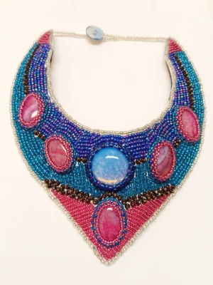 Färgglad pärlbroderad statement halsband - Unikt halsband med pärlbroderi i blått, rosa och lila. Halsbandet har en cool V-formad design och är täckt av glittrande pärlor och stora stenar i mitten. Perfekt för dig som vill sticka ut och älskar färg. Ingen stretch, knäpps med knapp i nacken.