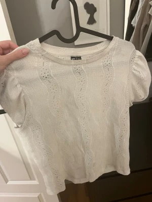 Vit blus med spets från Pieces - Säljer en söt vit blus från Pieces i storlek XS. Blusen har korta puffärmar och är dekorerad med vågiga spetsdetaljer och broderade hålmönster. Materialet känns lätt och luftigt, perfekt för varmare dagar. Rund halsringning och feminin stil.