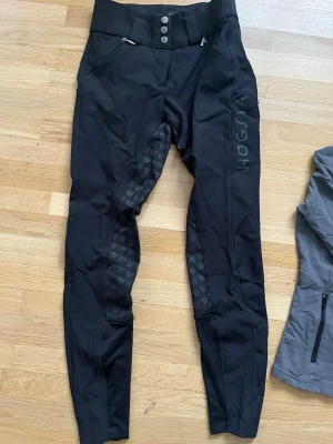 Svarta ridbyxor från Hogsta - Snygga svarta ridbyxor från Högsta med hög midja och tre knappar framtill. Byxorna har diskreta loggor längs benen och grepp i sätet för extra stöd. Två dragkedjefickor framtill och stretchigt material som ger skön passform. Dragkedja bak nedanför mudden. Knappt använda