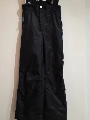 Svarta täckbyxor med reflexdetalj - Svarta täckbyxor med elastisk midja och hängslen. Utrustade med dragkedjor med blå detaljer och reflexband vid benslutet. Materialet är slitstarkt och passar perfekt för kalla dagar. Praktiska fickor med dragkedja och justerbara benslut.