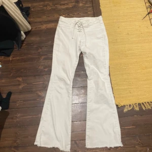 Vita bootcut byxor med snörning - Säljer ett par vita bootcut byxor med snörning i midjan och råa, fransiga benslut. Byxorna har hög midja och är tillverkade i ett mjukt bomullsmaterial. Perfekta för dig som gillar en retro och avslappnad stil.