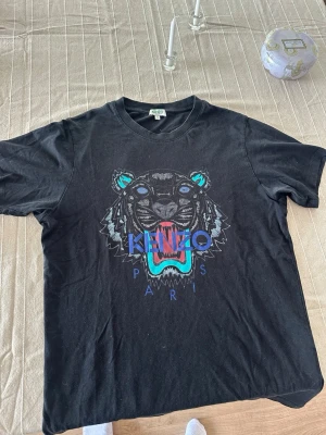 Svart Kenzo t-shirt med tigertryck - Säljer en svart t-shirt från Kenzo i storlek S med ikoniskt tigertryck på bröstet i blå, turkosa och röda detaljer. T-shirten har rund hals och korta ärmar, tillverkad i mjuk bomull. Perfekt för dig som gillar streetwear och vill sticka ut med ett coolt motiv.
