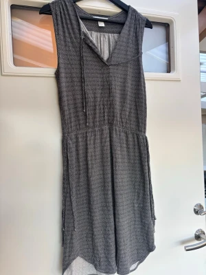 Svartvit mönstrad klänning med snörning - Svartvit ärmlös klänning från H&M med små cirkelmönster och v-ringad hals med snörning. Mjuk och luftig i bomullsmix, med resår i midjan för skön passform. Perfekt för dig som gillar avslappnad stil och snygga detaljer.