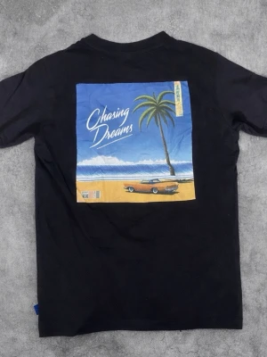 Svart t-shirt Chasing Dreams Only & Sons - Svart t-shirt från Only & Sons med trycket 'Chasing Dreams' på bröstet och en stor färgglad print på ryggen med strand, palm och bil. Klassisk passform och rund hals. Perfekt för dig som gillar streetstyle och snygg stl, Ny pris 249.
