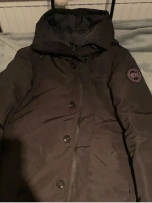 Canada Goose Chateau - Svart dunjacka från Canada Goose med huva och klassisk logga på ärmen. Jackan har knappar och dragkedja framtill, samt en stilren och enkel design. Perfekt för kalla dagar och har en skön, puffig känsla. Passar dig som vill ha både värme och stil. Pris kan diskuteras! 