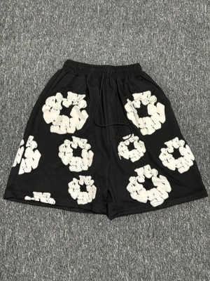 Svarta shorts med vit mönster - Säljer ett par svarta shorts med coola vita cirkelmönster över hela tyget. De har elastisk midja med snörning för skön passform och är tillverkade i ett mjukt syntetmaterial. Perfekta för dig som gillar streetwear och vill sticka ut.