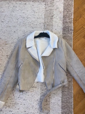 Beige bikerjacka från NLY Trend - Säljer en beige teddyjacka från NLY Trend i storlek 34. Jackan har mjukt foder i vitt teddy-material, bred krage, dragkedja framtill, två snedställda fickor med dragkedja och bälte i midjan. Perfekt för kalla dagar och riktigt snygg till jeans.