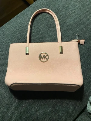 Ljusrosa handväska från Michael Kors - Säljer en ljusrosa handväska från Michael Kors i stilren design med guldfärgade detaljer och MK-logga framtill. Väskan har två rejäla handtag, en dragkedja upptill och en praktisk ytterficka med dragkedja på baksidan. Tillverkad i strukturerat skinn.