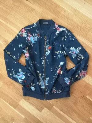 Blommig jacka från Vero Moda - Mörkblå jacka från Vero Moda med blommigt mönster i pastellfärger och dragkedja med rund ring. Jackan har ribbade muddar och klassisk bomberkrage.