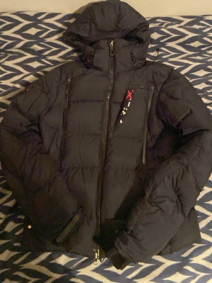 Svart pufferjacka från Ralph Lauren - Säljer en svart pufferjacka från Ralph Lauren med huva och dragkedja framtill. Jackan har en cool detalj med röd och vit brodyr på bröstet samt praktiska fickor. Perfekt för kalla dagar och riktigt skön passform.