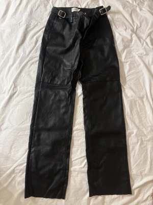 Svarta skinnbyxor med spännen - Säljer ett par svarta skinnbyxor från NAKD med raka ben och två coola spännen i midjan. Byxorna har klassisk femficksdesign och är tillverkade i mjukt skinnimitation. Perfekta för dig som vill ha en edgy look. Specifikt ifrån Josefine HJ NAKD kollektionen