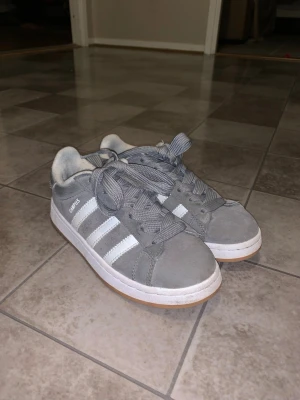 Grås Adidas Campus sneakers mocka - Säljer ett par grå Adidas Campus sneakers i mocka med klassiska vita ränder på sidorna och vit sula. Skorna har rund tå, snörning och gummisula i ljusbrun färg. Perfekta för dig som gillar retrostil och streetwear.