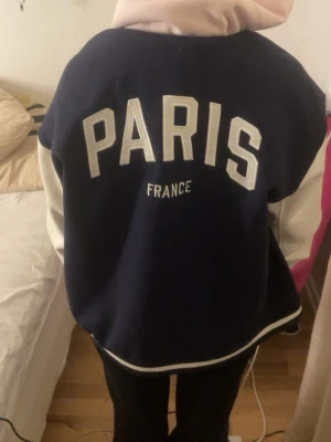 Mörkblå bomberjacka från H&M DIVIDED - Säljer en snygg mörkblå bomberjacka från H&M DIVIDED i storlek L. Jackan har vita ärmar, tryckknappar framtill och coolt PARIS-tryck på ryggen. Materialet är polyester och jackan har en loose passform med ribbade muddar vid ärmslut och nederkant.