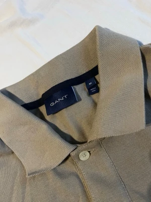 Beige pikétröja från GANT - Snygg beige pikétröja från GANT med klassisk krage och knappslå framtill. Tillverkad i mjuk bomull och har korta ärmar. Diskret GANT-logga broderad på bröstet. Perfekt för en clean och stilren look.