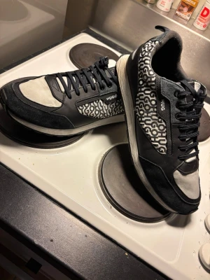 Svarta och vita sneakers från Hugo - Säljer ett par snygga sneakers från Hugo med svart och vit färg, coolt grafiskt mönster på sidorna och svarta snören. Skorna har rund tå och platt sula, tillverkade i en mix av skinn och syntetmaterial. Perfekta för dig som gillar streetstyle och vill sticka ut.