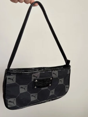 Svart axelväska från Puma - Cool svart axelväska från Puma med grått geometriskt mönster och loggor. Väskan har ett smalt axelband, detaljer i skinnimitation och dragkedja upptill. Insidan har ett fack med dragkedja för småsaker. Perfekt storlek för det viktigaste.