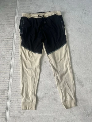 Beige och svarta mjukisbyxor C&A Active - Säljer ett par mjukisbyxor från C&A Active i storlek L. Byxorna är beige med svarta detaljer upptill och har snörning i midjan samt dragkedjor på fickorna. Materialet är mjukt och skönt, perfekt för chill eller träning. Snygg kontrast mellan färgerna och ribbade muddar vid bensluten.