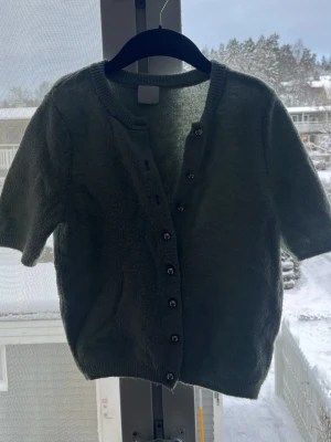 Grön kortärmad kofta med knappar - Mysig grön kofta med korta ärmar och rund hals. Koftan har knappar framtill och är gjord i ett mjukt, stickat material som känns fluffigt. Perfekt att slänga över en topp för en chill look.