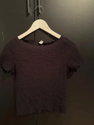 Svart ribbad topp med kort ärm - Säljer en svart topp med ribbad struktur och korta ärmar. Toppen har en rundad hals och är tillverkad i ett stretchigt material som sitter snyggt på kroppen. Perfekt att matcha med jeans eller kjol för en enkel och stilren look.