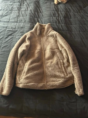 Beige teddyjacka från 157, XS - Mysig beige teddyjacka från 157 i storlek XS. Jackan har hög krage, dragkedja framtill och två fickor. Perfekt för kyliga dagar när du vill vara både varm och stilren. Materialet är fluffigt och ger en riktigt skön känsla.