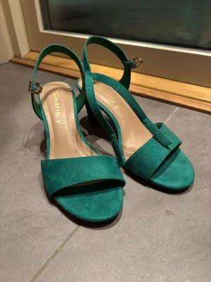 Gröna högklackade pumps med öppen tå, från Tamaris - Säljer ett par snygga gröna pumps med öppen tå, från Tamaris, i storlek 40. Skorna har en låg klack, öppen tå och justerbart spänne i guld. Tillverkade i mocka med mjuk innersula för extra komfort. Perfekt för dig som gillar färg och vill sticka ut.