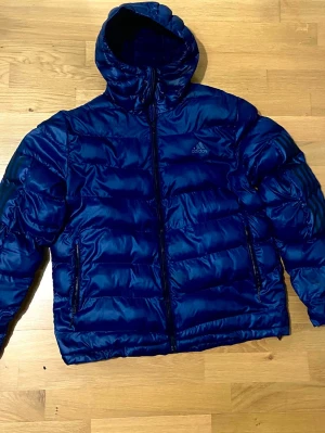Blå pufferjacka från Adidas, storlek L - Blå pufferjacka från Adidas med huva och dragkedja framtill. Jackan har två sidofickor med dragkedja och ett diskret Adidas-logga på bröstet. Materialet känns lätt, men också perfekt för kyliga dagar. Passformen är normal och jackan är fodrad för extra värme.