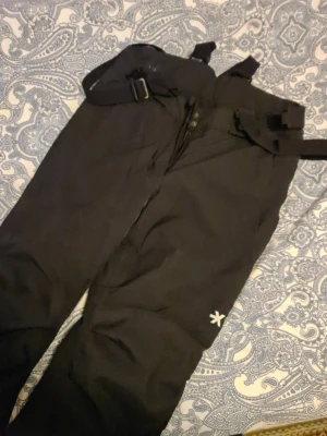 Svarta skid-täckbyxor från Everest 164 - Svarta skid-täckbyxor från Everest med justerbara hängslen och bältesdetaljer. Byxorna har en enkel design med logga på benet och är gjorda i slitstarkt syntetmaterial som passar perfekt för kalla dagar utomhus. Snygga och praktiska för vinteraktiviteter. Cirka använt en gång p.g.a för liten storlek