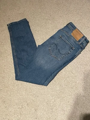 Blå jeans från Jack & Jones - Snygga blå jeans från Jack & Jones med klassisk femficksdesign och läderpatch bak i midjan. Jeansen har en rak passform med normal midja och är tillverkade i slitstarkt bomullsmaterial. Perfekta för dig som gillar enkel och tidlös stil.