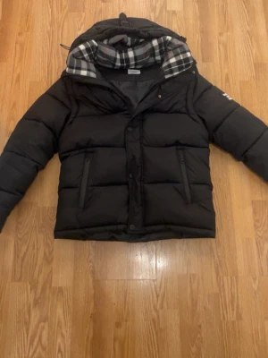 Svart pufferjacka från Burberry S - Svart pufferjacka från Burberry med klassisk rutmönstrad huva och tydlig logga på ärmen. Jackan har dragkedja och knappar framtill samt två fickor med dragkedja. Materialet är polyester och jackan har en loose passform.