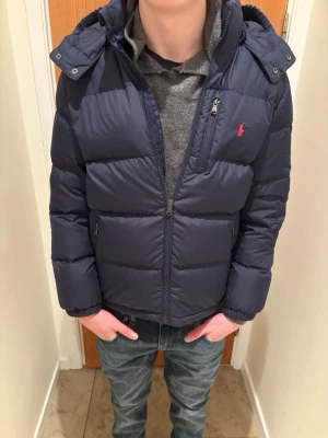 Mörkblå pufferjacka från Ralph Lauren - Säljer en mörkblå pufferjacka från Ralph Lauren med huva och dragkedja framtill. Jackan har en röd logga på bröstet, flera fickor med dragkedja och är riktigt skön för kalla dagar. Perfekt för dig som vill ha en stilren och varm jacka med snygg passform.