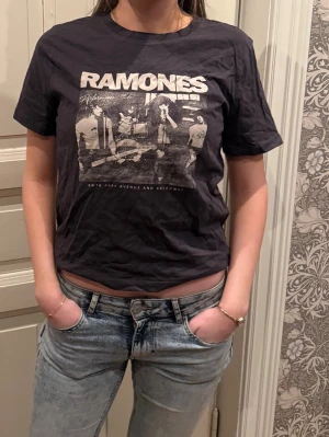 Svart Ramones t-shirt med tryck - Säljer en svart t-shirt med Ramones-tryck framtill. Klassisk passform med korta ärmar och rund hals. Perfekt för dig som gillar bandtröjor och vill ha en cool, avslappnad stil. Materialet känns som bomull och trycket är i vitt och grått.