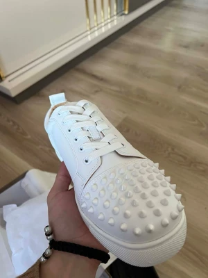 Vita sneakers från Christian Louboutin - Säljer ett par riktigt snygga vita sneakers från Christian Louboutin. Skorna är i skinn och har coola nitar över tån, samt klassisk snörning. Perfekta för dig som vill sticka ut med en lyxig och edgy vibe.