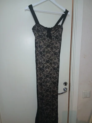 Lång svart spetsklänning med blommönster - Säljer en elegant långklänning med breda axelband och svart spets med blommigt mönster över beige tyg. Klänningen har en söt snörning framtill vid bysten och figurnära passform. Perfekt för dig som vill sticka ut med en klassisk men ändå unik look.