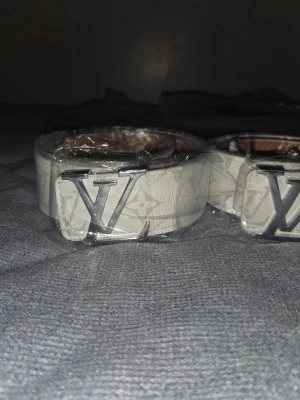 Vit Louis Vuitton bälte med LV-spänne - Snyggt vitt bälte från Louis Vuitton med klassiskt LV-spänne i silverfärg. Bältet har diskret mönster med logotyp och är tillverkat i skinn. Perfekt för att lyfta din outfit med en lyxig touch.