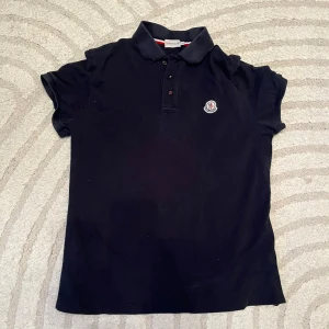 Svart Moncler piké topp - Säljer en svart piké topp från Moncler i storlek L. Toppen har klassisk krage, korta ärmar och knappar framtill. På bröstet sitter Monclers broderade logotyp. Materialet är mjuk bomull som känns skönt mot huden.