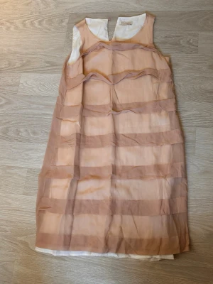 Beige ärmlös kortklänning - Säljer en beige ärmlös kortklänning från Designer Remix med transparenta horisontella ränder och v-ringad hals. Klänningen har ett lager av tunn, skir polyester och ett vitt innerfoder. Perfekt för dig som gillar stilrena detaljer.