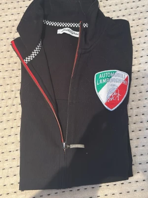 Svart Lamborghini vindjacka - Svart vindjacka från Lamborghini med coolt broderat märke på bröstet i grönt, vitt och rött. Jackan har dragkedja med röd detalj och schackrutigt band vid kragen. Perfekt för dig som gillar motorsport och snygg streetstyle.