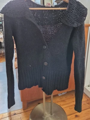 Svart stickad kofta från Indiska - Svart stickad kofta från Indiska i storlek M. Koftan har en bred, grovstickad krage och tre stora knappar framtill. Långärmad med ribbad nederkant och muddar. Perfekt för lager-på-lager och chill stil.