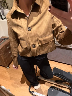 Cropped mockajacka från ZARA - Säljer en snygg croppad jacka från ZARA i mjuk mocka-look. Jackan är brun med stora fickor framtill, klassisk krage och knäppning med två knappar. Perfekt för dig som gillar stilrena och trendiga plagg med lite edge.