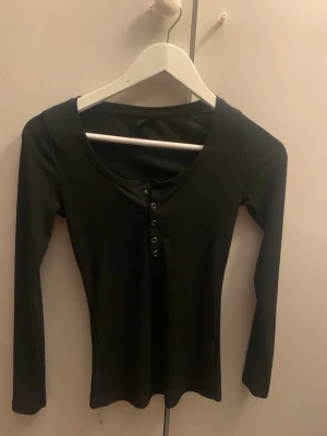 Svart ribbad långärmad topp från SHEIN - Svart långärmad topp från SHEIN i storlek M. Toppen är ribbad och har en rund halsringning samt knappar framtill. Materialet är mjukt och stretchigt, vilket gör den bekväm att bära. Perfekt att styla med jeans eller kjol för en enkel look. Jag tycker att det är storlek S en av knapparna finns tyvärr inte
