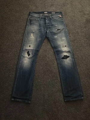 Replay jeans med slitningar, blå 31/32 - Säljer ett par blå Replay Waitom jeans med coola slitningar och hål på både fram- och baksida. Klassisk femficksmodell med raka ben och tydlig tvättad look. Genomgående hål på knäna. Märkesdetaljer syns vid fickan och bak på linningen. Materialet är jeans i bomull.